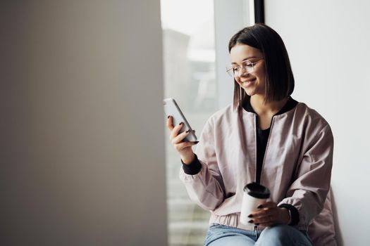 Young Woman in Glasses Drinking Coffee and Using Smartphone While Sitting on Windowsill