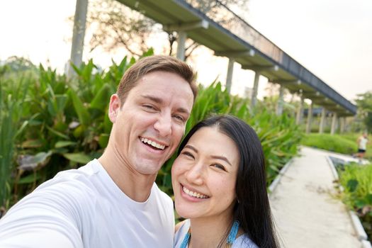 Portrait of a multiethnic couple laughing while taking a selfie in an urban park during sunset