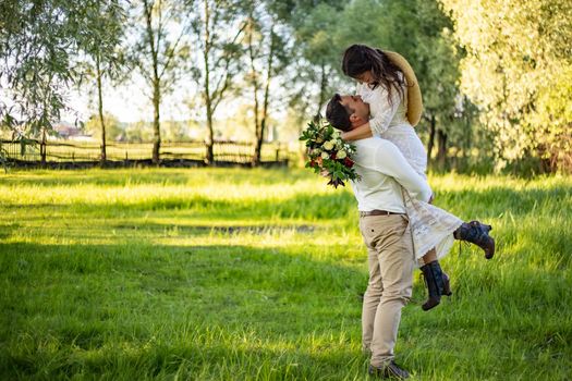 Stylish happy bride, groom hold picked up in hands and dancing. The groom raised and spins bride. Newlyweds kissing. On background on nature in the courtyard of house. Close up. Wedding first dance