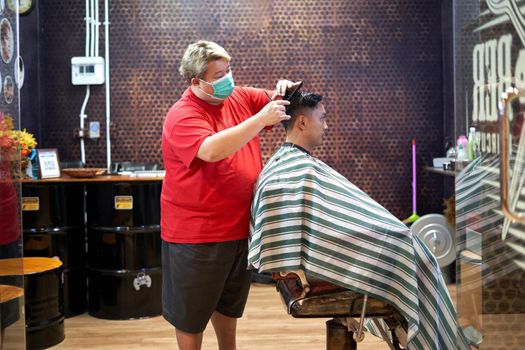 Obese barber wearing mask standing cutting the hair of an asian homosexual client in a barber shop