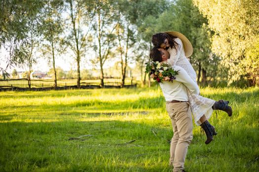 Stylish happy bride, groom hold picked up in hands and dancing. The groom raised and spins bride. Newlyweds kissing. On background on nature in the courtyard of house. Close up. Wedding first dance