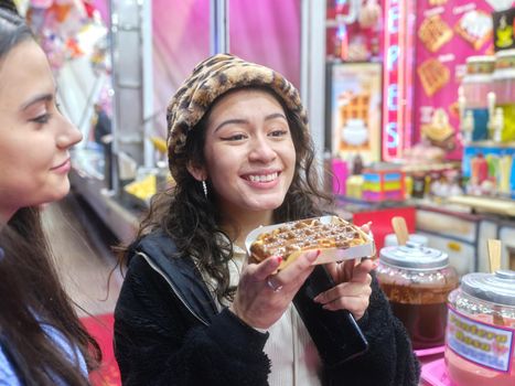 Smiling latina woman in hat eating a chocolate waffle in a night fair next to a friend