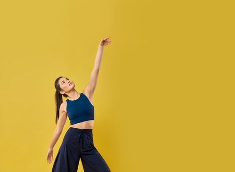 Relaxed female dancer looking up, raising hand in spacious studio. Front view of stylish girl with ponytail moving arms gracefully while dancing against orange studio background. Concept of lifestyle.