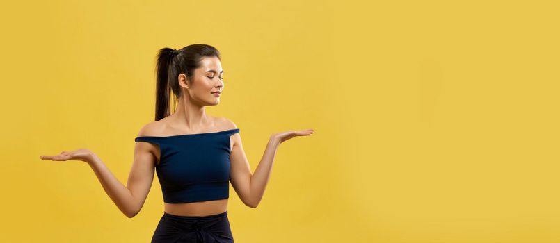 Smiling woman standing on knees, keeping hands palms up indoor. Portrait view of graceful girl in black top kneeling with arms bent at elbows, isolated on orange studio background. Concept of posing.