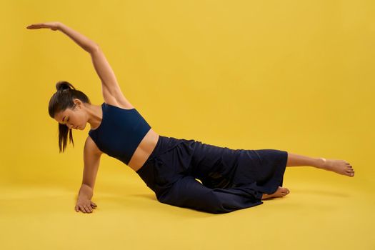 Slender brunette girl practicing pose of yoga, stretching on floor, isolated on yellow studio background. Front view of attractive woman in activewear exercising alone indoor. Concept of yoga asana.