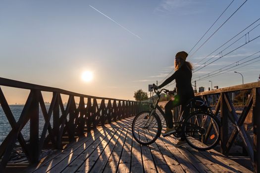 Silhouette of young girl with curly hair. Riding a bicycle on wooden bridge at sunset on the promenade road of Maresme, Catalonia, Spain.