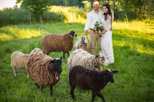 Stylishly dressed handsome groom with a beard in black sunglasses hugs wife. Bride in a dress with a bouquet puts a hat on and laughs. Rustic wedding in the style of boho at the ranch