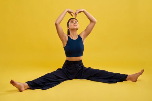 Female yoga coach doing split, stretching muscles during exercise indoor. Front view of slim girl sitting in twine, holding arms over head during yoga practice, isolated on yellow. Concept of yoga.