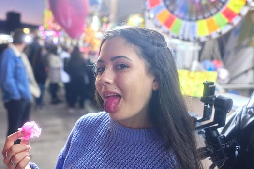 Latina woman showing her pink tongue while eating cotton candy in a crowded night fair