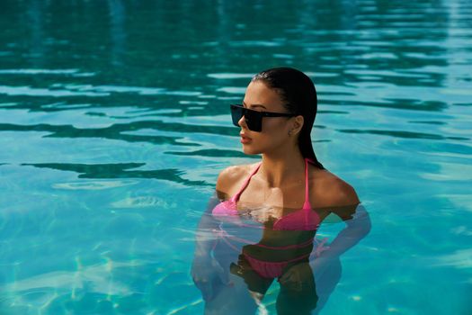 Front view of pretty young female with long wet hair swimming in pool. Seductive girl relaxing, having vacation, enjoying, sunbathing in sunglasses. Concept of summertime.