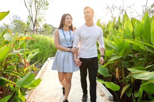 Multicultural couple walking hand in hand on a path surrounded by tropical plants in an urban park.