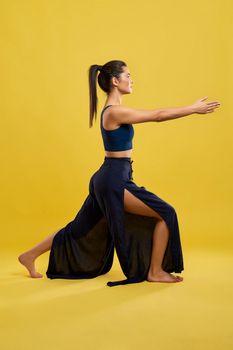 Yogi girl with ponytail keeping arms stretched, while staying in yoga pose indoor. Side view of fit woman in wide pants, practicing yoga, isolated on orange studio background. Concept of yoga.