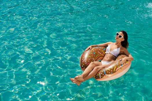 Above view of slim young female having rest in summer outdoors. Brunette girl wearing white swimsuit and sunglasses, lying on donut, swimming, sunbathing. Concept of youth.