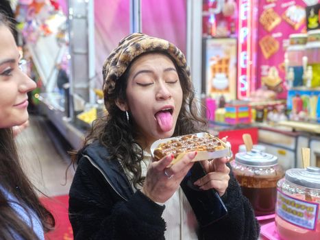 Latina young woman joking with the tongue out before eating a chocolate waffle in an evening fair