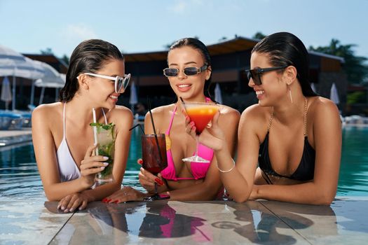 Front view of three pretty girls standing in swimming pool. Seductive young females wearing swimsuits, holding cocktails, relaxing, looking at each other. Concept of summer.