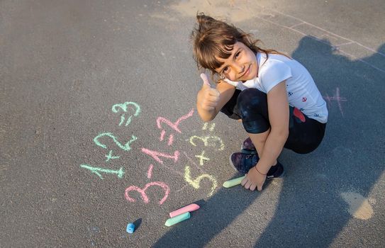 The child solves equations on the asphalt with chalk. Selective focus. Kid.