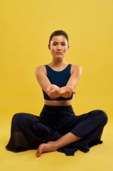 Pretty woman sitting in easy pose of yoga, stretching arms forward indoors. Front view of young blurred girl holding hands palms up, isolated on orange studio background. Concept of gesturing.