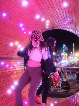 Vertical photo of two young fashion women dancing inside a tube of an attraction at a night fair