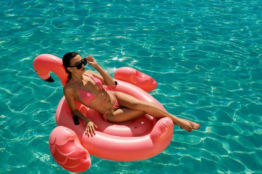 Above view of slim brunette girl swimming in pool. Pretty young female wearing pink swimsuit and sunglasses, lying on pink flamingo, sunbathing. Concept of summertime and youth.