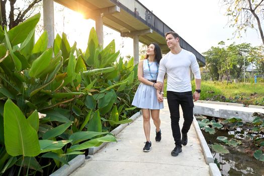 Multiethnic couple walking distracted for a path of an urban park with tropical plants during sunset
