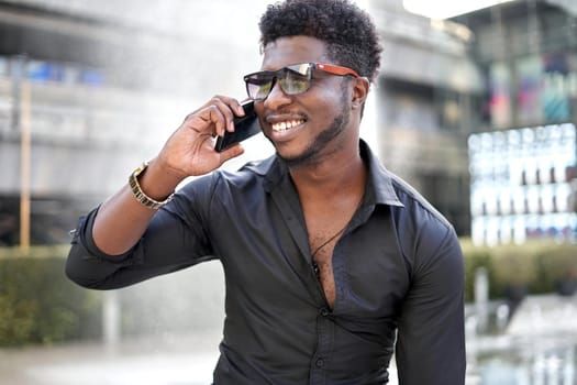 Portrait of a smiling black stylish man talking to the mobile phone outside a shopping mall