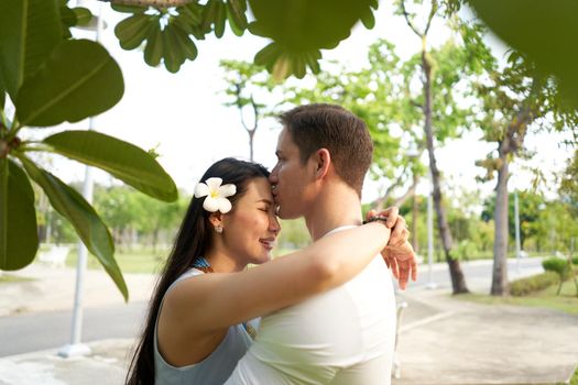 South african man kissing the forehead of a thai woman with a flower in the hair in a park
