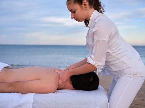 Side view of a young chiromassage therapist giving a shoulder massage to young man on a beach in Valencia.