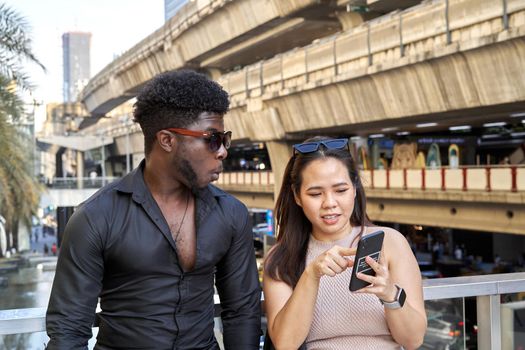 Asian woman showing information of the screen of a mobile phone to a afro friend in a shopping mall