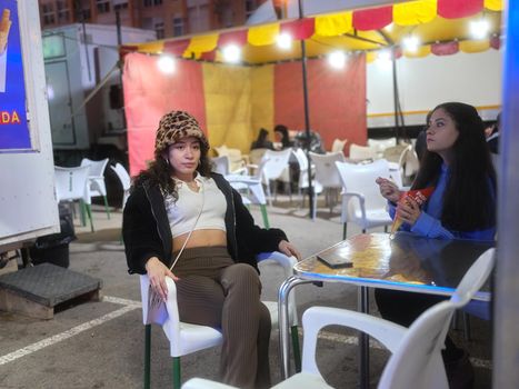 Two latina women sitting on a food stall on a night fair while eating french fries