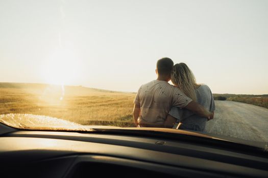 Back View Through Front Car Window on Young Couple Hugging Near the Hood, Woman and Man Enjoying Road Trip at Sunset