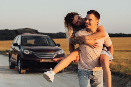 Caucasian Man Carries Girlfriend on His Back on Background of Their SUV Car, Young Couple Enjoying Their Road Trip at Sunset