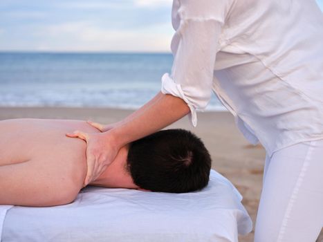 Side view of a young man receiving shoulder massage from a female chiromassage therapist on a beach in Valencia.