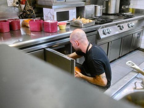 Chef crouching while storing food in stainless steel kitchen fridges in the kitchen of a restaurant