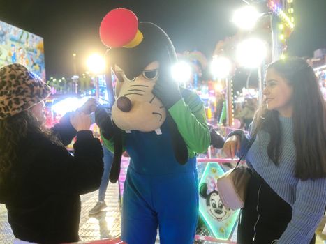 Two latina women playing with an actor dressed like Goofy dog in a theme park at night