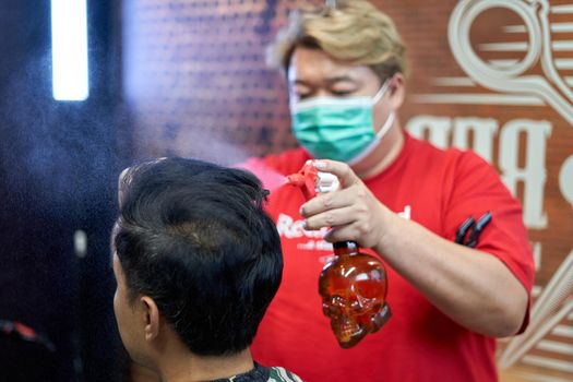 Asian fat barber with mask wetting a customer's hair with a dispenserto cut it in a barber shop