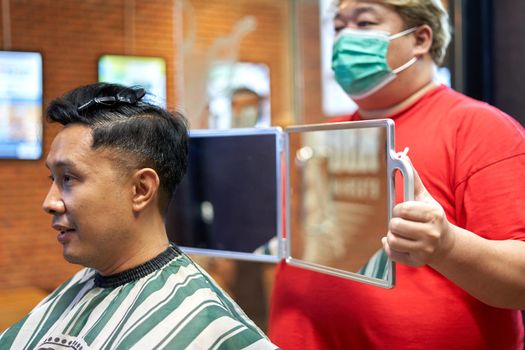 Fat barber with mask displaying the back of the head to a gay customer using a mirror in a barber shop