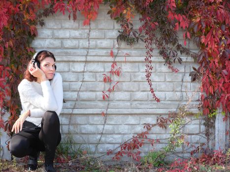 Woman crouching down listening to music with mobile next to a wall with climbing plants in autumn