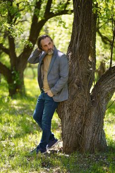 A nice looking mature birded man in casual standing under tree on the grass looking behind the tree with hands in pockets. Freelancer spend time working outdoors.