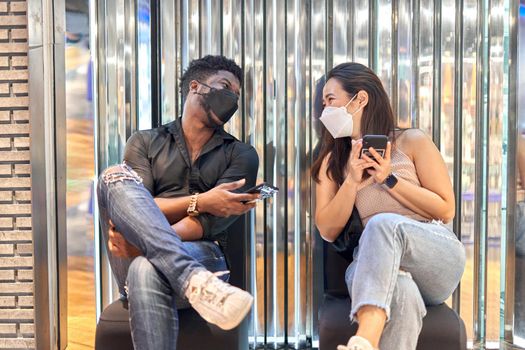 Two multiracial smiling friends wearing mask using the mobile while sitting on a shopping mall
