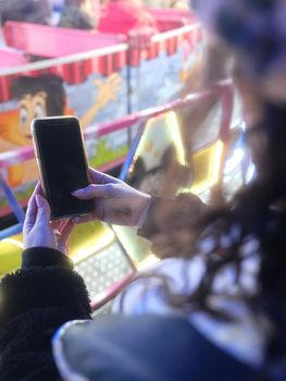 Vertical photo of the hands of a Woman using the mobile in front of a night fair