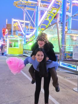 Female friend carrying other in the back in the middle of a night fair while holding a cotton candy