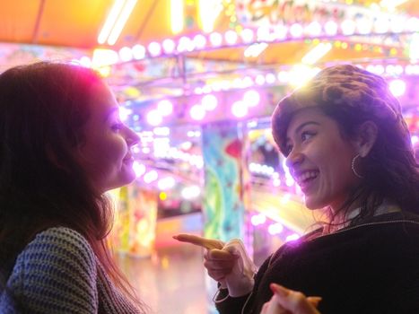 Happy latina woman pointing to another while smiling next to an attraction at night fair