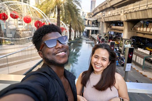Back man and asian woman smiling to the camera while taking a selfie outside a shopping mall