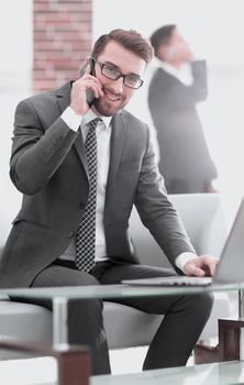 Smiling man talking on mobile phone while using laptop computer at desk in study.