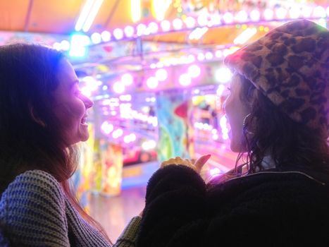 Two latina women chatting and smiling in front of an attraction of a colorful evening fair