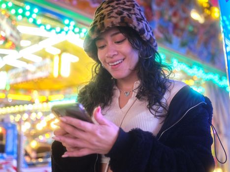 Latina woman in leopard hat smiling while using the mobile next to a fairground attraction at night