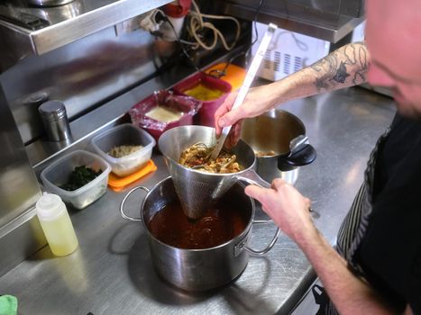 Chef strains food using a strainer in a pot to prepare a tasting menu in the kitchen of a restaurant