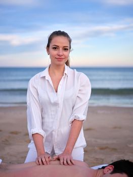 Vertical image of a young therapist performing a chiromassage treatment on a young man while looking at camera outdoors on a beach in Valencia.