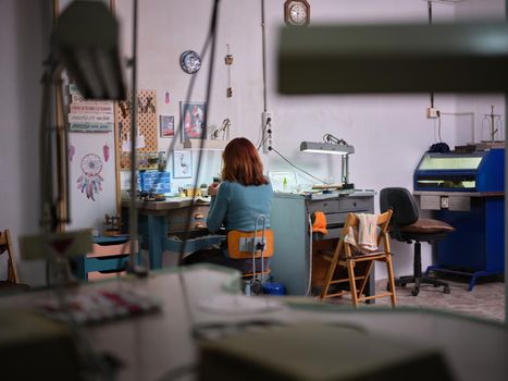 Rear view of an adult worker woman working in her jewelry artisan workshop sitting on workbench.