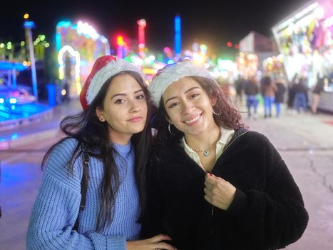 Portrait of two beauty latina women wearing christmas hat while looking at the camera in a night fair